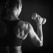 Eine Person mit einem geflochtenen Zopf steht im Profil und hält eine Hantel in der erhobenen rechten Hand. Der Hintergrund ist dunkel und kontrastreich.