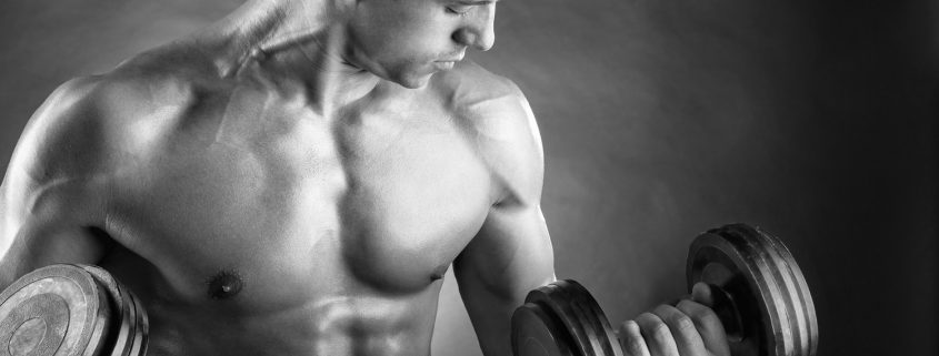Ein muskulöser junger Mann hebt eine Hantel, während er nachdenklich seinen Blick senkt. Das Bild ist in Schwarz-Weiß gehalten.