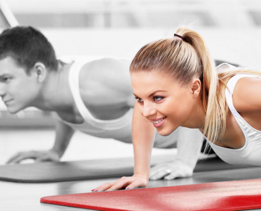 Eine Frau und ein Mann machen gemeinsam Liegestütze auf Yogamatten. Die Frau lächelt, während der Mann konzentriert aussieht.