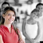 Eine lächelnde Frau steht im Vordergrund, während verschwommene Menschen im Hintergrund in Schwarz-Weiß sichtbar sind. Die Frau trägt ein rotes Oberteil.