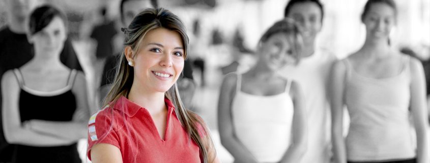 Eine lächelnde Frau steht im Vordergrund, während verschwommene Menschen im Hintergrund in Schwarz-Weiß sichtbar sind. Die Frau trägt ein rotes Oberteil.