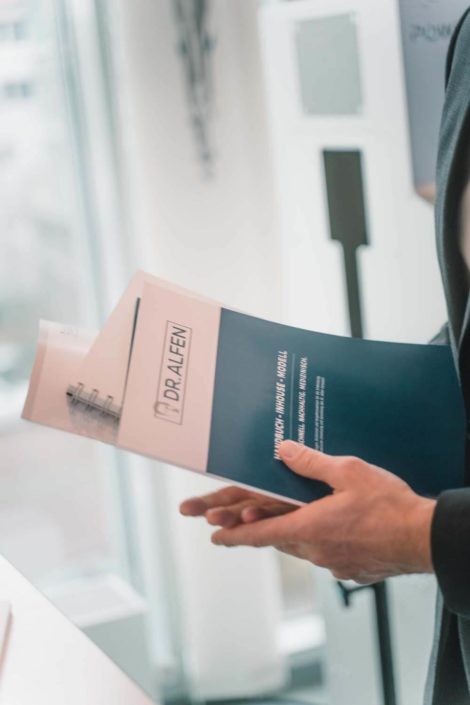 Eine Person hält ein aufgeschlagenes Dokument mit dem Titel "Dr. Alfen" und einem blauen Cover in der Hand, während sie in einer hellen Umgebung steht.