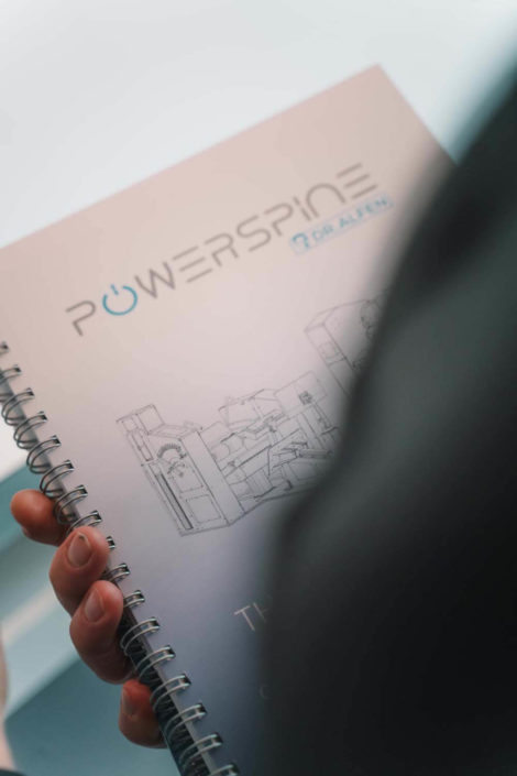 Eine Hand hält ein Spiralbuch mit dem Titel „POWERSPINE“, das technische Zeichnungen zeigt. Der Hintergrund ist unscharf.