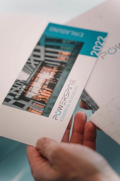 Eine Hand hält ein Informationsblatt zum POWERSPINE Kongress in Würzburg, das ein modernes Gebäude im Hintergrund zeigt sowie das Jahr 2022.