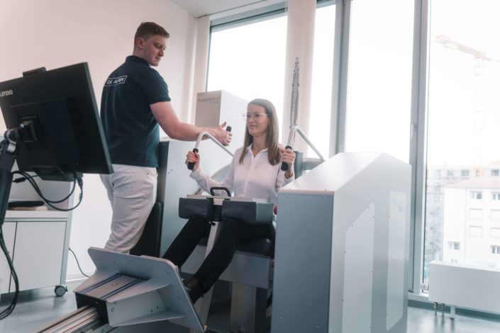 Ein Physiotherapeut unterstützt eine Patientin, die auf einem modernen Trainingsgerät sitzt, während sie die Griffe in den Händen hält. Der Raum ist hell und modern eingerichtet.