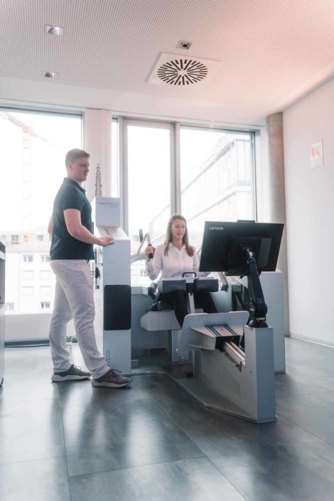 In einem modernen Raum stehen ein Mann und eine Frau. Die Frau sitzt an einem Trainingsgerät mit Monitor, der Mann beobachtet sie aufmerksam.