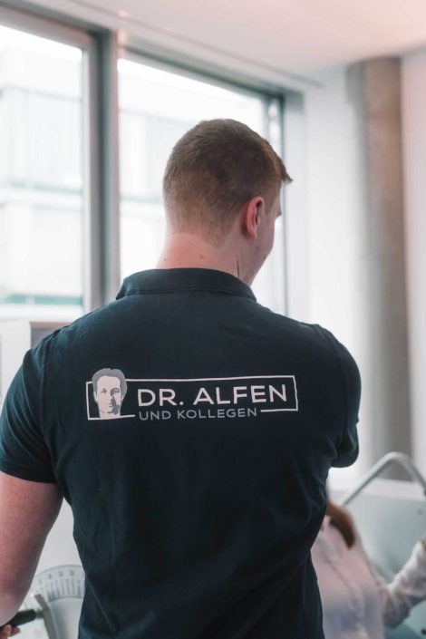 Ein Mann mit kurzer Frisur trägt ein schwarzes Poloshirt mit dem Aufdruck "Dr. Alfen und Kollegen" auf dem Rücken. Im Hintergrund sind Fenster und eine Person im weißen Kittel zu sehen.