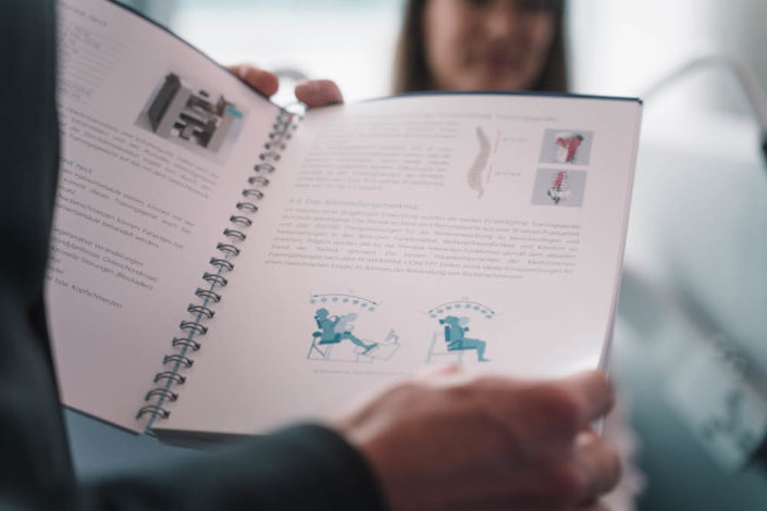 Eine Person hält ein geöffnetes Heft, das Informationen und Illustrationen zu ergonomischen Sitzpositionen und Haltungsrichtlinien enthält. Im Hintergrund ist eine andere Person unscharf zu sehen.