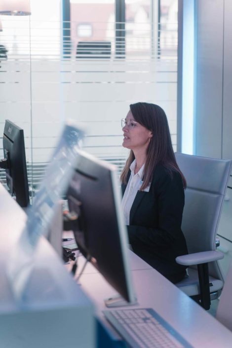 Eine Frau in einem Anzug sitzt an einem modernen Schreibtisch vor zwei Computern und spricht, während sie auf den Bildschirm schaut. Hintergrund mit Fenstern.
