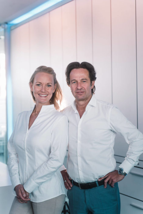 Ein Mann und eine Frau stehen lächelnd nebeneinander in eleganter, heller Kleidung vor einer modernen, hell erleuchteten Büro-Umgebung.